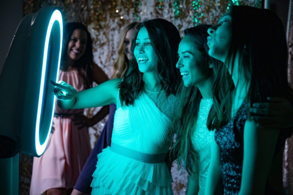 Drie vrouwen staan bij een photobooth, lachend en poserend voor de camera. Ze dragen feestelijke outfits en lijken volop te genieten van het moment.